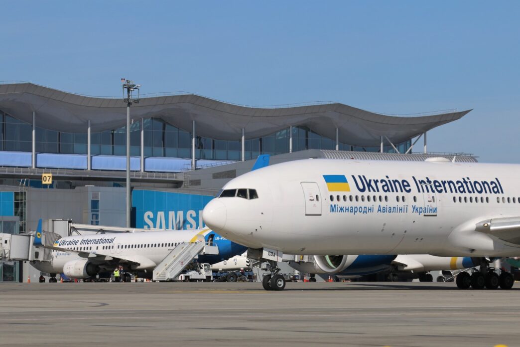 Широкофюзеляжний літак із написом Ukraine International стоїть біля скляного термінала аеропорту, поруч інші літаки та наземна техніка