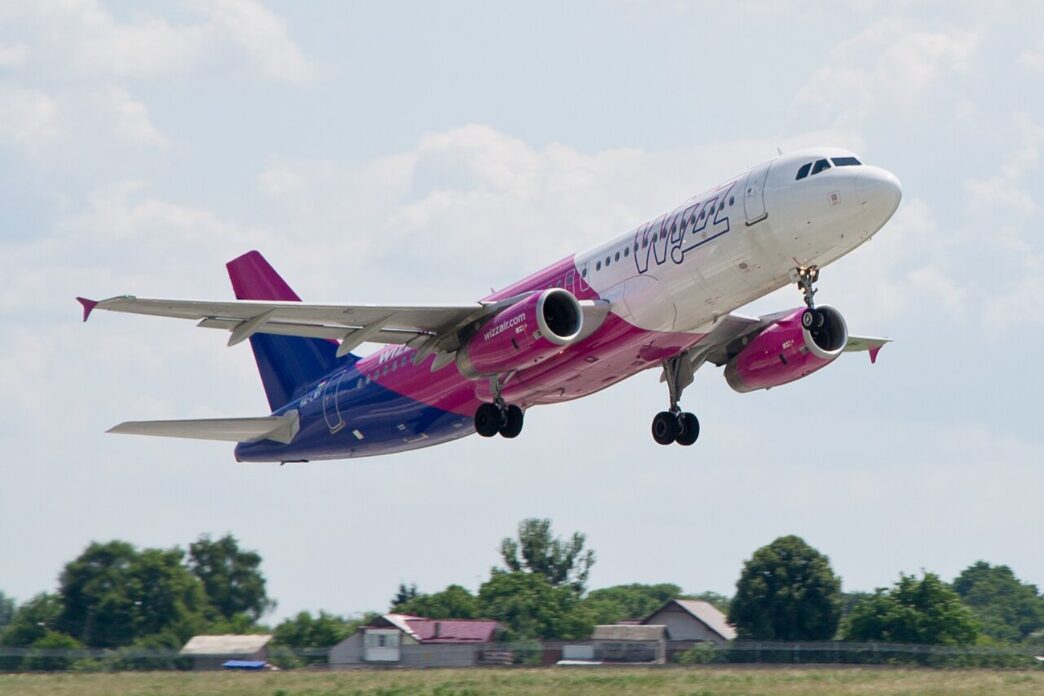 Пасажирський літак Wizz Air у фіолетово-рожевій лівреї набирає висоту після зльоту, під ним зелені поля та будинки