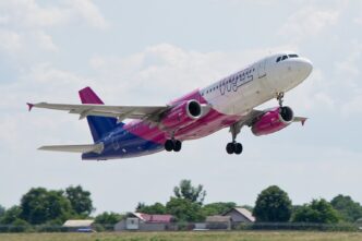 Пасажирський літак Wizz Air у фіолетово-рожевій лівреї набирає висоту після зльоту, під ним зелені поля та будинки