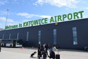 Пасажири з валізами йдуть уздовж темно-синьої будівлі терміналу з великим зеленим написом Welcome to Katowice Airport, ліворуч стоїть міський автобус