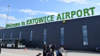 Пасажири з валізами йдуть уздовж темно-синьої будівлі терміналу з великим зеленим написом Welcome to Katowice Airport, ліворуч стоїть міський автобус