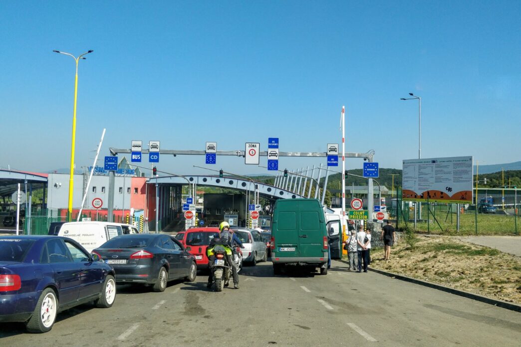 Черга легкових автомобілів і мікроавтобусів перед пунктом пропуску на автомобільному кордоні, біля узбіччя йдуть пішоходи, сонячний день