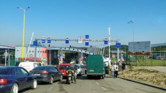 Черга легкових автомобілів і мікроавтобусів перед пунктом пропуску на автомобільному кордоні, біля узбіччя йдуть пішоходи, сонячний день