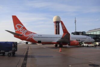Пасажирський літак Boeing 737 у червоно-білій ліврі SkyUp на пероні аеропорту, поруч наземна техніка, на задньому плані диспетчерська вежа і термінал, денне світло