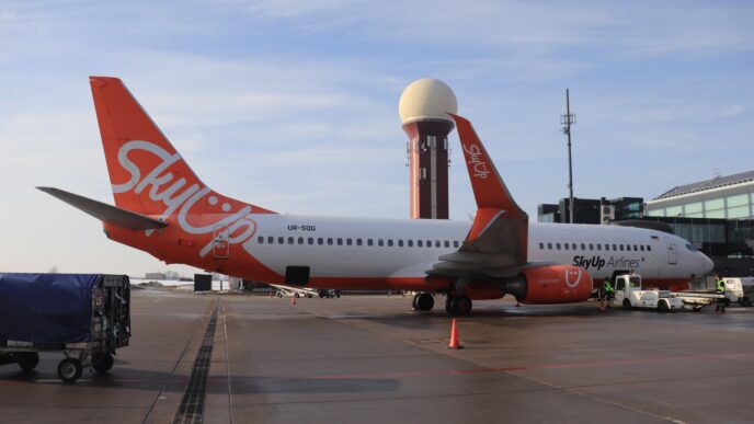 Пасажирський літак Boeing 737 у червоно-білій ліврі SkyUp на пероні аеропорту, поруч наземна техніка, на задньому плані диспетчерська вежа і термінал, денне світло